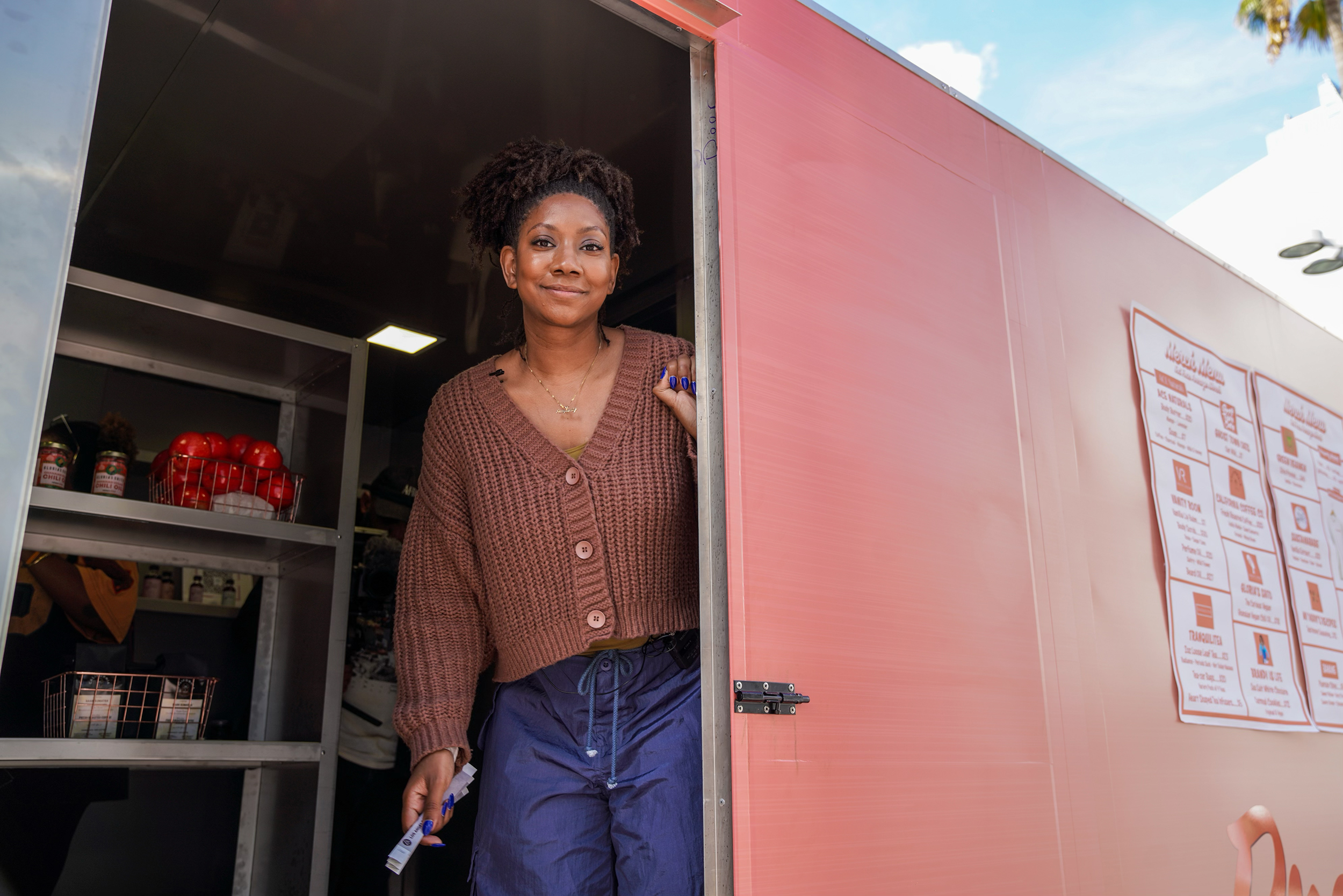A Black woman poses inside the doorway of a pink trailer with a shelving unit behind her.