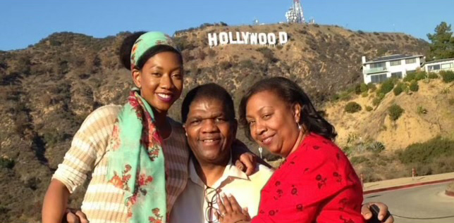 A young Black woman with a green headscarf stands smiling with her Black parents in front of the California Hills with the Hollywood sign in the background.
