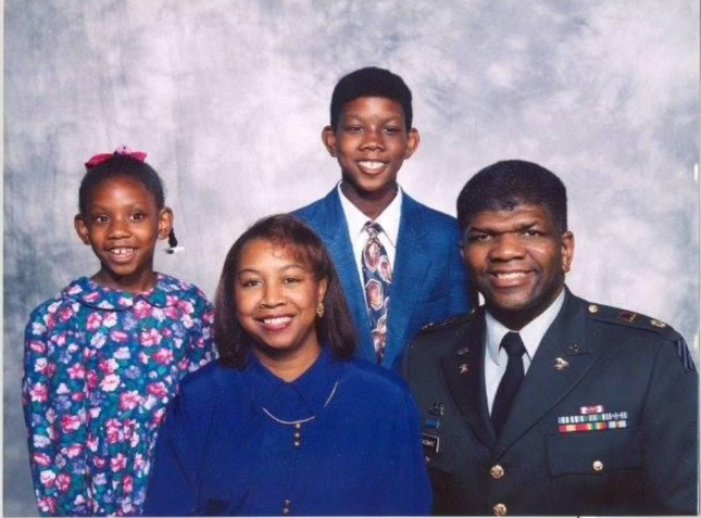 A Black family of four poses for a portrait. A Black uniformed military personnel is situated on the right side of the frame.
