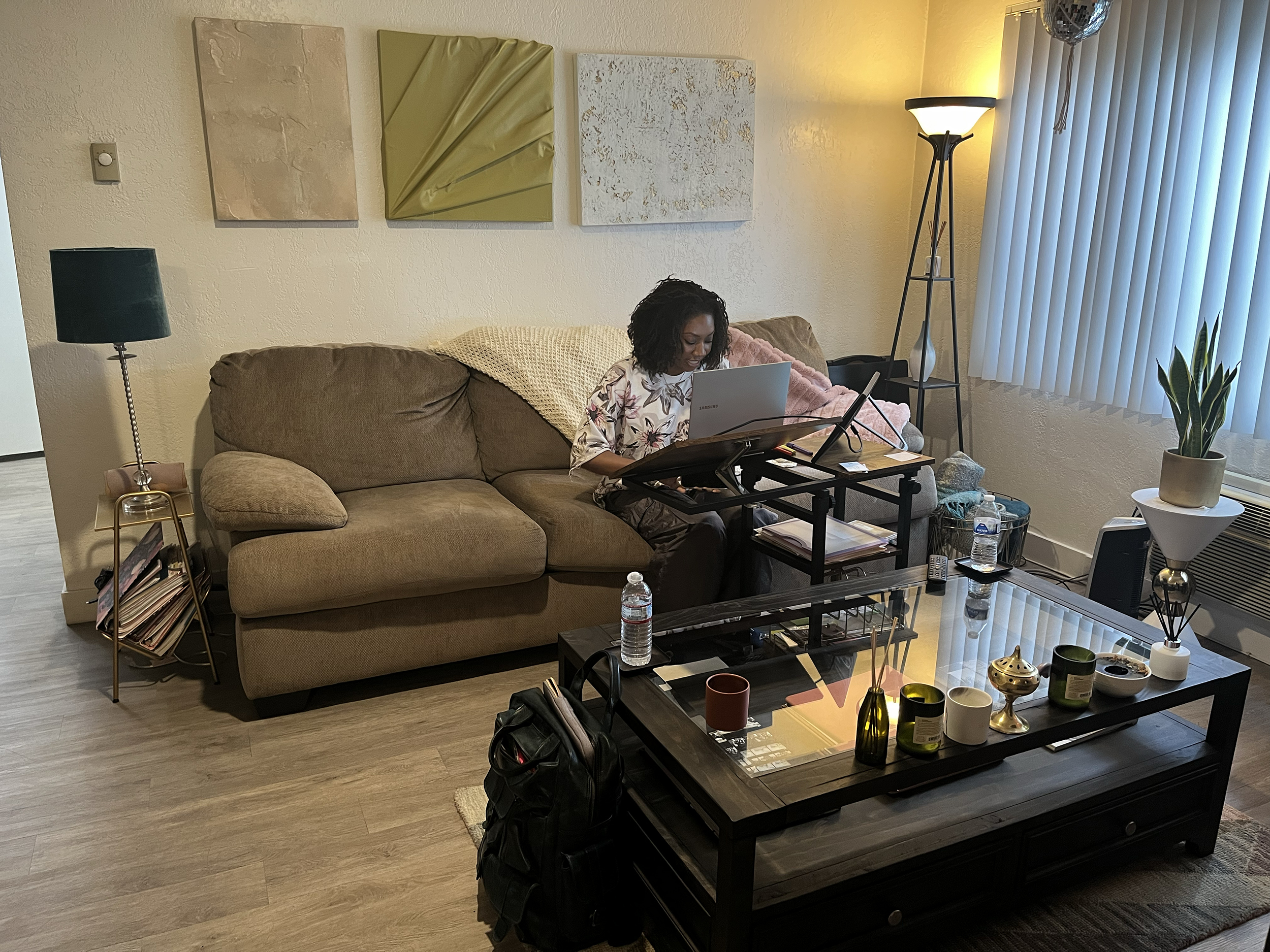 A Black woman sits on a brown couch with her laptop in front of her on a computer stand.