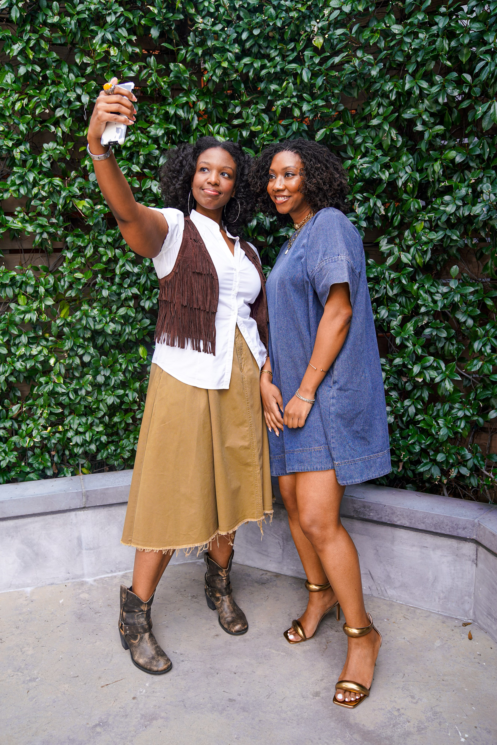Two Black women are pictured in front of a green backdrop taking a selfie together with a cellphone.