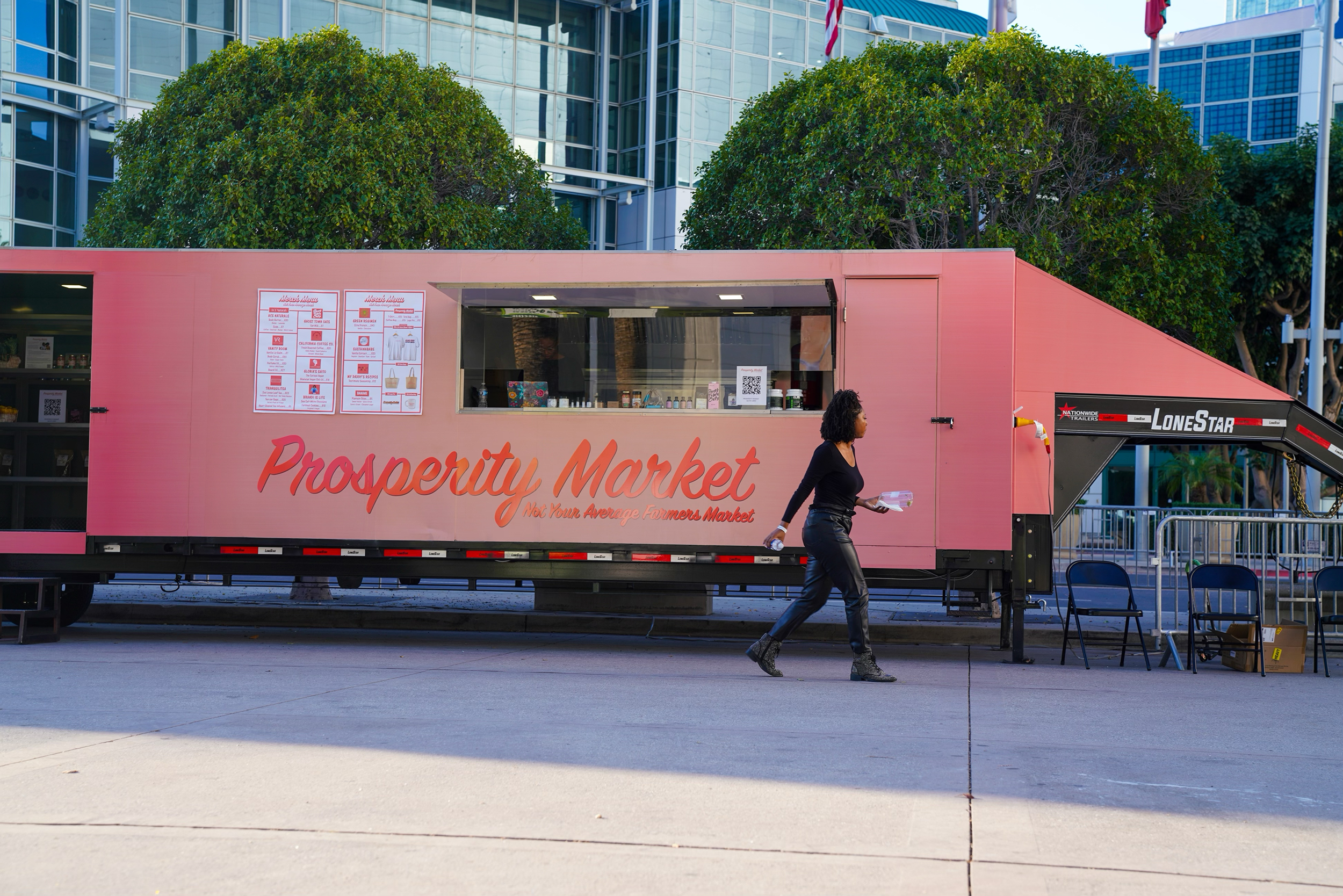 A Black woman walks in front of a pink mobile trailer which has the words “Prosperity Market” written alongside the bottom half of the trailer.