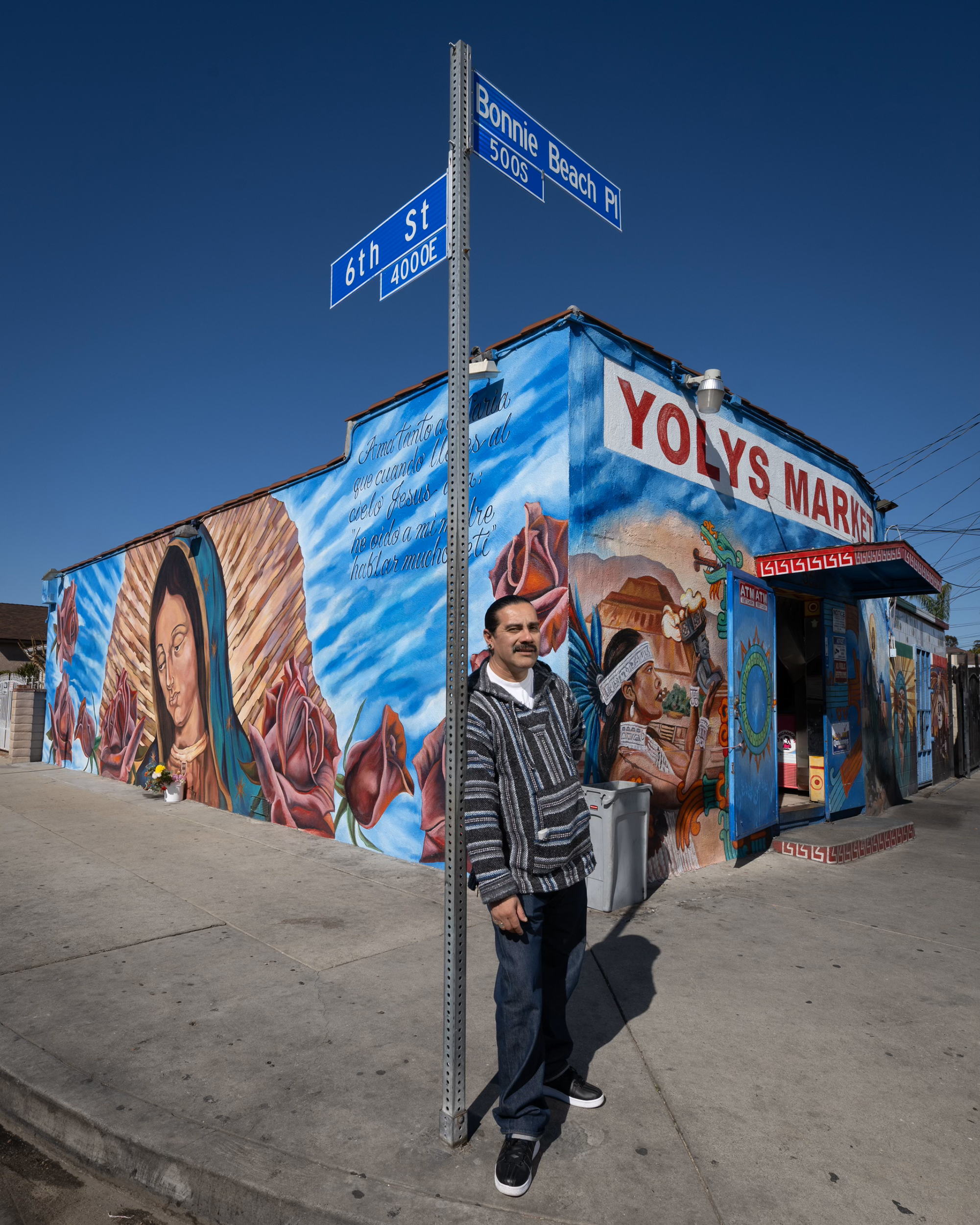 A Latino man wearing a black and gray-striped sweater stands at a street post at the corner of 6th St. and Bonnie Beach Place. Behind him, a colorful mural of the Virgin of Guadalupe and an Aztec person wrap around the walls of a building labeled “Yolys Market.”