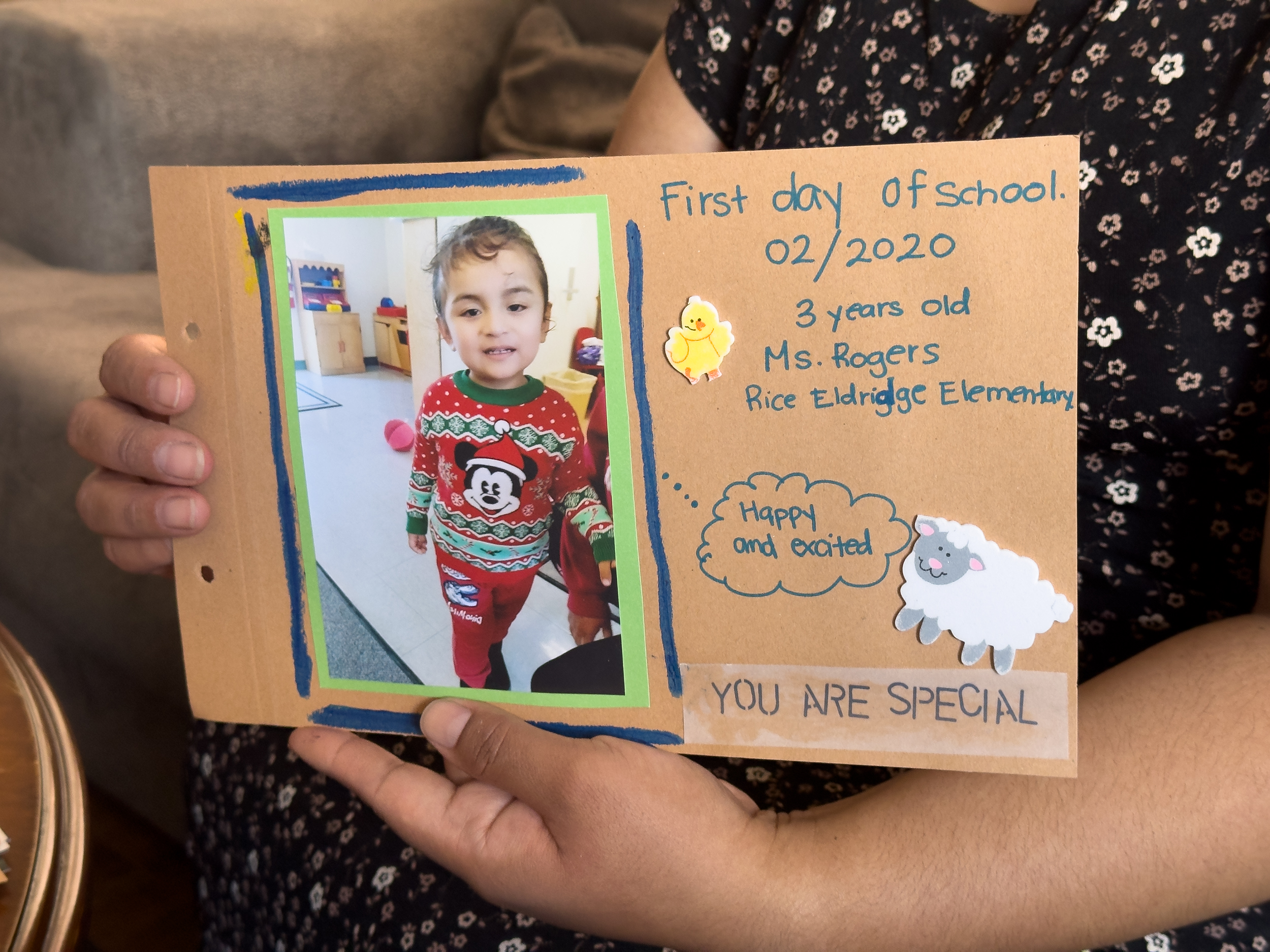 A woman holds a photograph of her son on his first day of preschool. The photo is mounted on a piece of cardboard with cutouts of a chicken and a sheep and the words, “You are special.”