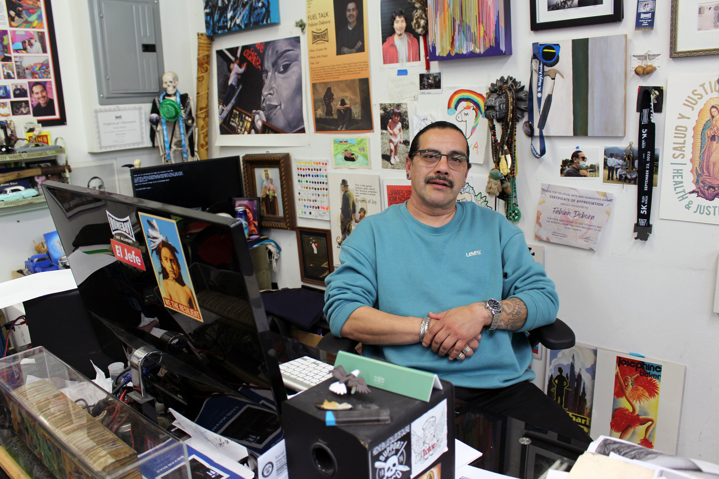 A Latino man with a mustache and glasses is wearing a teal Levi’s sweatshirt. He is sitting at a desk with art covering the walls behind him.