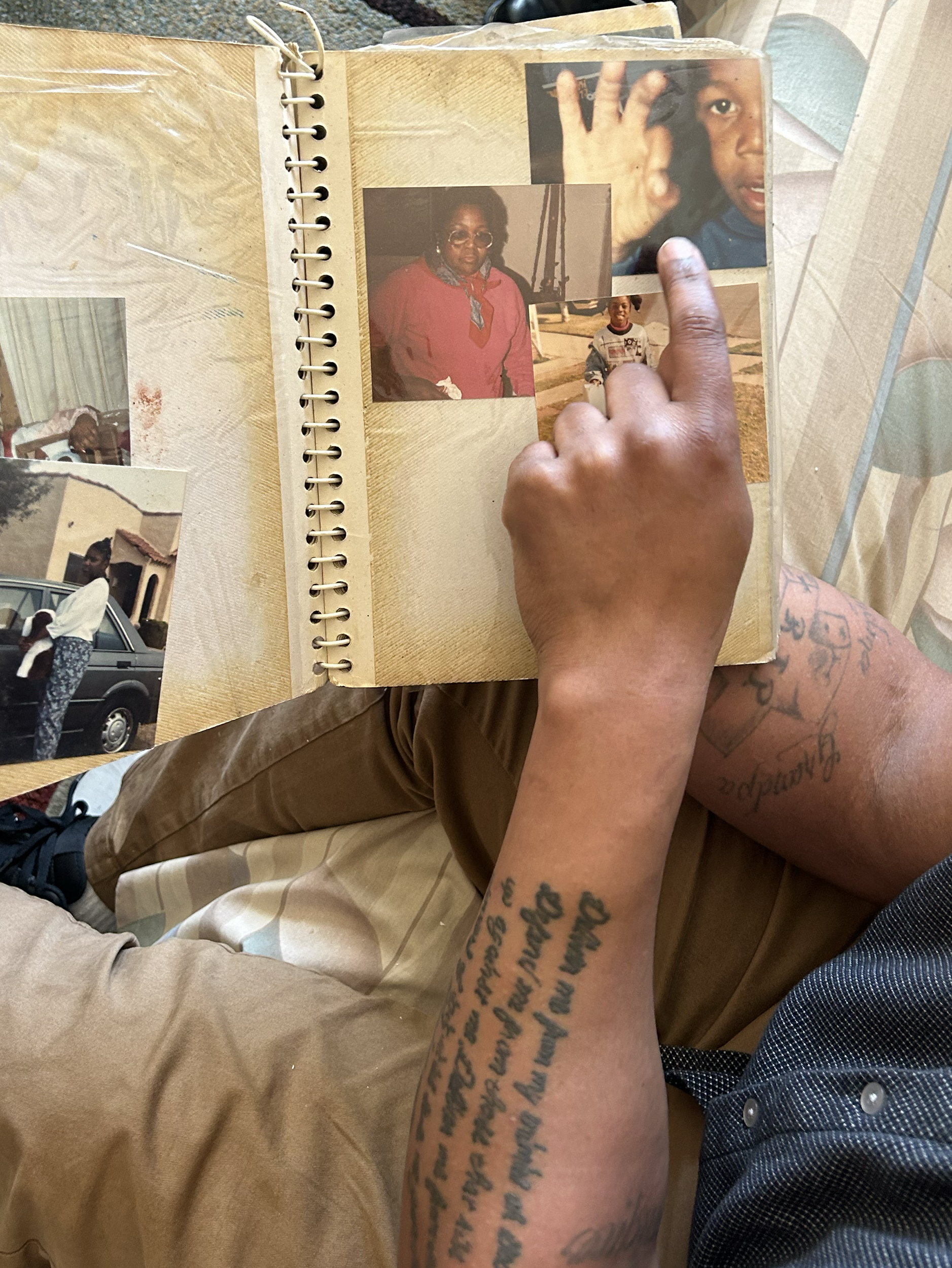 A black man’s tattooed arm hovers over a photo album as his hand points to a photo of a Black child (himself) making signs with his fingers.