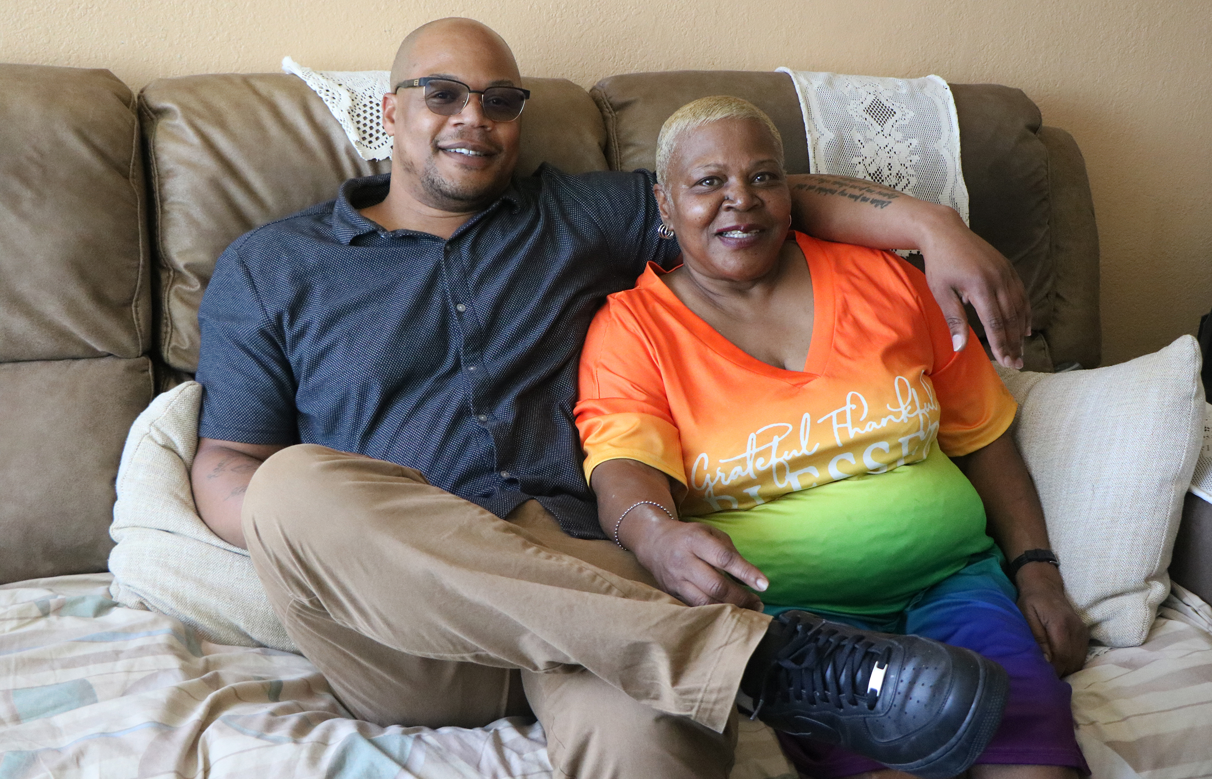Black man (right) sits on a couch with his arm around his Black mother. The two are smiling.